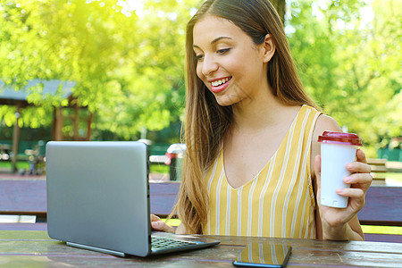 拿着杯子的女孩手持笔记本电脑的年轻女商务人士 拿着咖啡坐在公园里背景
