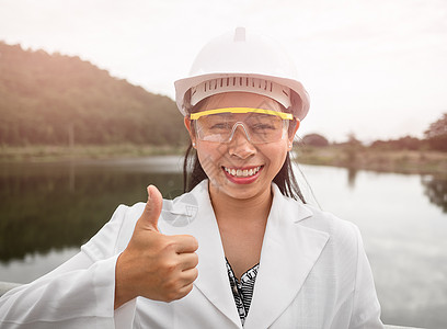 身戴头盔的女工程师对着镜头微笑 同时在水坝建筑工地拿着一个带有大拇指的数字平板板来生产电能图片