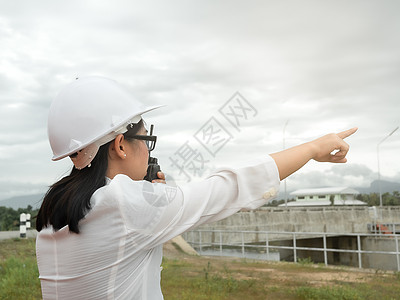 水电工程师戴着白色头盔的女工程师在大坝施工现场使用无线电与建筑工人通信发电背景