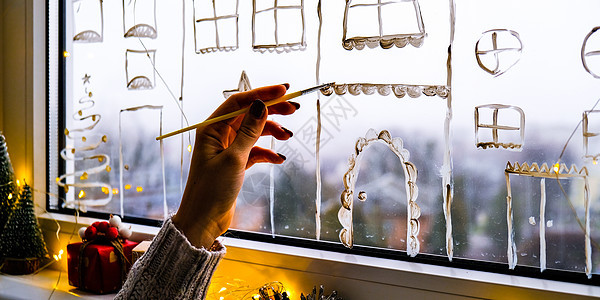 女手在窗户玻璃上画圣诞假期装饰 新年风景 城市建筑花环灯 Hygge 装饰和圣诞节概念建筑学花环风格传统假期地标展示雪片庆典反思图片