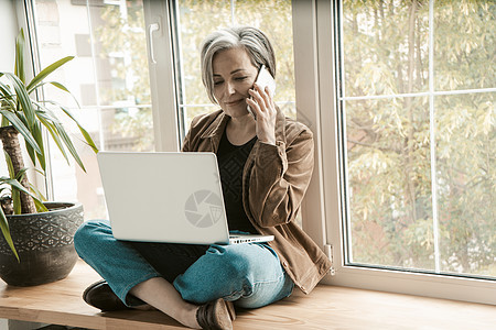 在笔记本电脑上工作时谈手机的迷人女人 头发花白的老妇人盘腿坐在宽阔的窗台上 背靠着窗户 着色图像图片