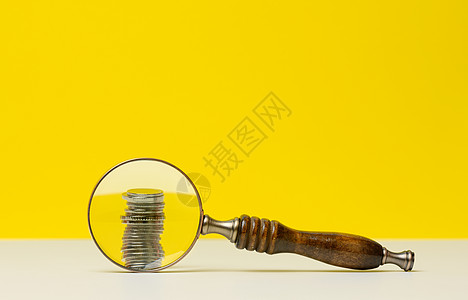 一堆硬币和一个木制放大镜在白色桌子上 黄色背景 预算资金分析 收支控制的概念 微薄的薪水 为创业公司筹款图片