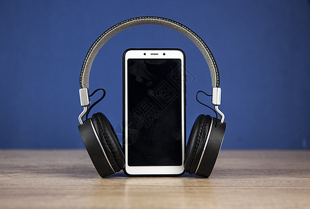 木桌上的黑色耳机和智能手机与蓝色墙纸背景技术电缆工具电话宏观金属屏幕商业立体声音乐图片