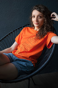 一个身穿亮橙色 T 恤的漂亮卷发女孩的女性肖像 她坐在家里的公寓里 背景是黑暗的 快乐的人的概念姿势深色眼睛工作室冒充学生成人橙图片