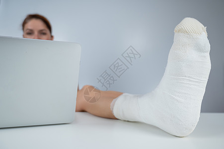 白种女人用石膏抬起腿去工作台 在白色背景的笔记本电脑上工作假期笔记绷带援助保健经理互联网写作助手疾病图片