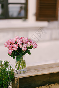餐桌招待会 一束粉红玫瑰花束装在玻璃透明 有水的花瓶中 站在一个木盒子上 背景是开着窗门的窗户图片