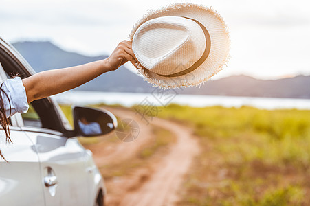 快乐的女人手拿着帽子在开窗车外 有草地和高山湖泊背景 人们在假期的公路旅行中放松作为旅行者的生活方式 运输和旅行概念图片