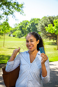 在公园外 一位身穿蓝领衬衫 面带微笑的混血非洲裔美国女性举着一张带有微芯片的蓝色塑料信用卡 她的棕色钱包斜挎在肩上图片