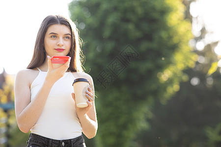 在公园散步的女孩用智能手机说话 快乐的女孩使用智能手机语音识别音频 ai 消息语音功能在线行走在公园 她手里拿着一杯咖啡图片