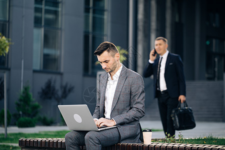 穿着西装坐在长凳上用笔记本电脑工作的男人 远程工作概念 远程工作 穿着西装的孤立男人 在户外笔记本电脑上打字的商务人士图片