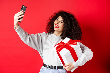 节假日和技术概念 快乐的女士带着礼物自拍 拿着现时手机和智能手机 站在红色背景上广告购物女孩电话黑发生活情感女性工作室卷曲图片