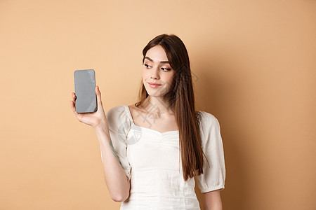 有吸引力的年轻女性展示空智能手机屏幕 微笑和看一眼 站在蜜蜂背景上站着工作室购物女士情感假期伙伴学生电话技术褐色图片