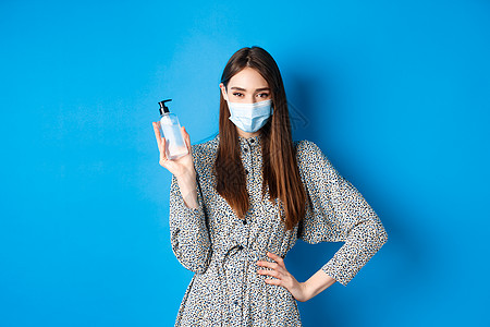 Covid19 社会不稳定和保健概念 穿着医疗面具的美丽女孩使用手防血清 拿着抗食药瓶 站在蓝色背景上女孩情感工作室女士微笑幸福图片