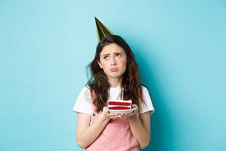 节日和庆祝活动 戴着派对帽的悲伤女孩拿着生日蛋糕 带着深思熟虑的心烦意乱的表情看着别处 在她的生日那天感到孤独和喜怒无常 站在蓝图片