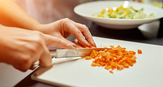 女人切成小立方块 在一块木板胡萝卜上 为了沙拉 它站在一个盘子里的背景图片