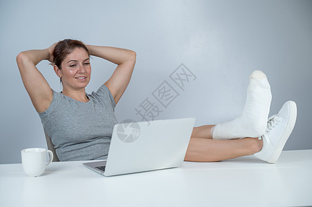 白种女人用石膏抬起腿去工作台 在白色背景的笔记本电脑上工作经理外科保健疾病事故假期援助写作创伤助手图片