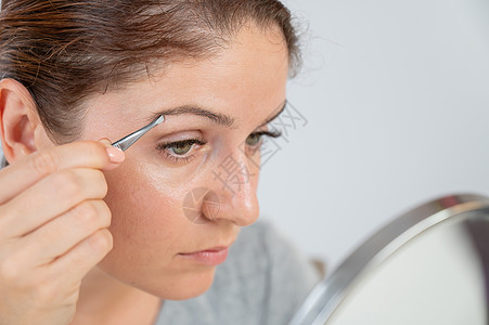 白种女人看着镜子 用tweezers自己做眉毛校正 然后用tweezers喜悦眼睛睫毛程序投标头发疼痛闲暇治疗护理图片