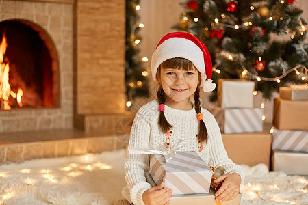 美丽微笑的小女孩带着礼物坐在地板上看着相机 穿着白色套头衫和红色帽子 孩子在壁炉和圣诞树旁摆姿势图片
