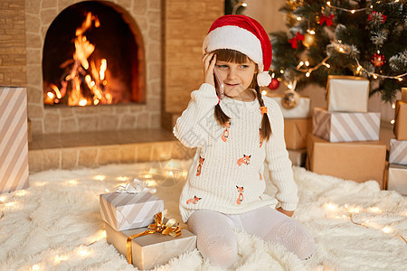 新年圣诞树前 穿着漂亮的白色毛衣和红色圣诞帽的快乐小女孩微笑着 摆姿势 拿着智能手机靠近耳朵 坐在礼盒旁 说话 感谢礼物图片