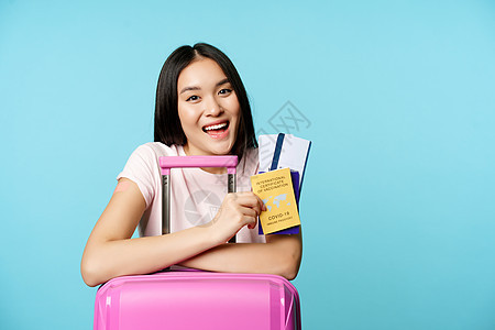 充满热情的亚裔女孩出示了护照和健康证明19岁 微笑高兴 站立着西装 蓝底脸蛋女性文档证书假期旅行女士接种航程乘客卫生图片