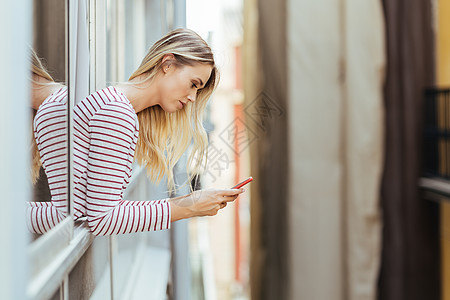 迷人的女人靠着智能手机 从她家的窗户向外倾斜图片