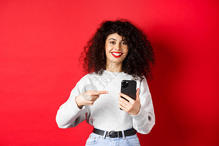 带着智能手机 微笑满意 推荐应用程序 站在红色背景下站立的漂亮女士 她满脸欢欣鼓舞工作室电话技术黑发女性生活广告促销女孩情感图片