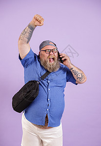 兴奋的肥胖男人 用袋式谈话 关于智能手机提高拳头 在紫底背景图片