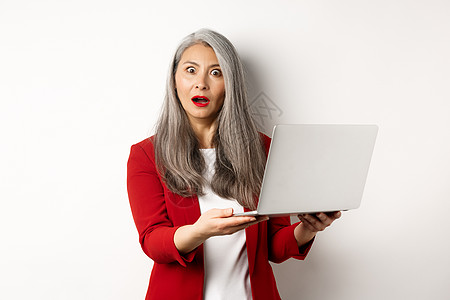 令人印象深刻的亚洲老年妇女在笔记本电脑上工作 盯着照相机惊讶地看着镜头 站在白色背景旁站着办公室祖母企业家商务化妆品成功电子商务背景图片