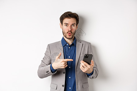 令人兴奋的西装男说哇 在智能手机上找到一些东西 指着触目惊心的手机 白色背景成人男性技术成功套装促销办公室企业家商务生活图片
