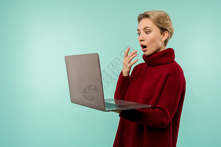 穿着毛衣的快乐金发美女 看着笔记本电脑屏幕 张着蓝背景的张开嘴图片