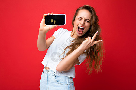快乐微笑的年轻金发女性漂亮 穿着白色 T 恤站在红色背景中孤立 复制空间拿着手机 手拿智能手机 空屏幕显示用于模拟看着相机并展示图片