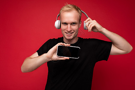 身穿黑色 T 恤和白色耳机的微笑快乐英俊的金发年轻人孤立地站在红色背景上 复制空间拿着智能手机 展示手机屏幕听音乐 看着相机玩得图片
