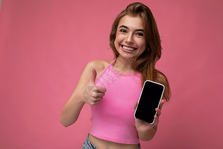 身穿粉红色上衣的美丽快乐的年轻金发女性 在粉红色背景中与世隔绝 手握空荡荡的空间 展示带有空显示屏的手机 用于模拟看着相机并竖起图片