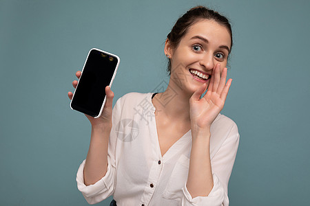 美丽积极的年轻金发卷发女性穿着休闲白衬衫 与世隔绝在蓝色背景墙上 拿着手机 展示带有空屏幕的智能手机 用于模拟看着相机和说秘密幸图片