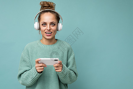 特写照片 美丽积极快乐的年轻女性穿着蓝色毛衣 在蓝色背景中被隔离 戴着白色蓝牙无线耳机 听音乐 使用手机在互联网上在线交流 看着图片