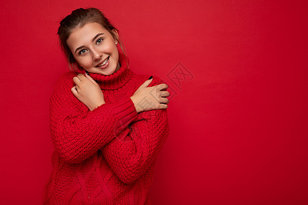 穿着温暖的红色毛衣的迷人可爱微笑的年轻女性被隔离在红色背景墙上 手放在胸前看着相机头发拥抱空间女士图片