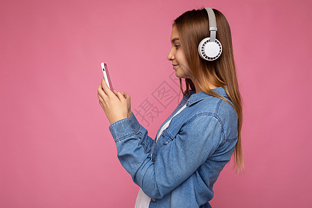 侧面特写照片 美丽的年轻女子穿着时尚休闲装 与彩色背景墙隔开 戴着白色无线耳机 听音乐 使用手机短信查看智能手机显示屏音乐播放器背景图片