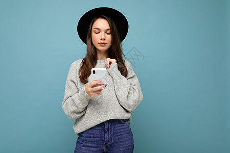 穿着黑帽子和灰色毛衣的美丽的年轻黑发美女 拿着智能手机看着在背景上孤立的电话简讯图片