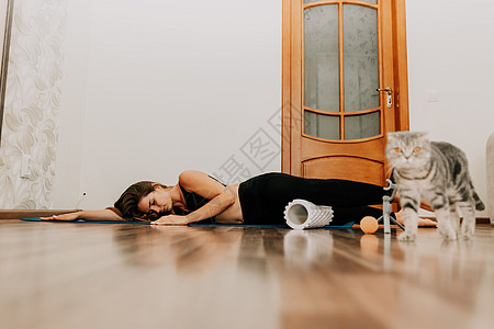 留着长头发的年轻女性 穿着黑色运动服的健身教练 在家里和猫一起在瑜伽垫上做伸展运动和普拉提 繁殖英国折耳猫 女性健身瑜伽常规概念图片
