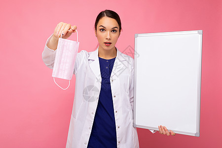 身穿白色医疗大衣的女医生用空白板 有复制文字空间和防护面罩 粉红色背景隔离在纸上流行概念预防药品黑发护士女士面具小样感染木板女性图片