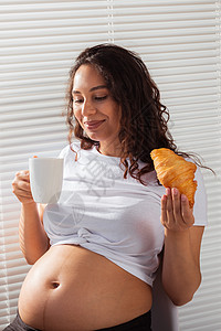 孕妇饮食快乐怀孕年轻美丽的女人在早餐期间吃羊角面包 愉快的早晨概念和怀孕期间的积极态度产妇女性产假幸福微笑孩子食物母亲烘烤糕点背景