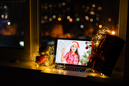 笔记本电脑站在桌子上 上面放着礼物和圣诞帽 在舒适的房间里 小女孩在屏幕上微笑 祝圣诞快乐 新年快乐图片
