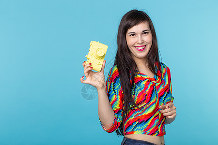 微笑的年轻女子手里拿着一个黄色的老式相机模型 在蓝色背景上摆姿势 有复制空间 业余和专业摄影的概念女孩闲暇摄影师潮人女性衣服快乐图片