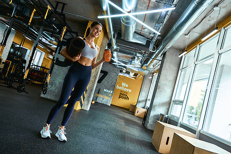 一位身穿运动服的年轻漂亮健身女性拿着瑜伽垫和一瓶水 在健身房锻炼时看着窗户微笑图片