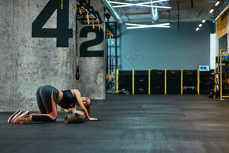 一位身穿运动服 用泡沫滚筒锻炼 在工业健身房的瑜伽垫上做伸展运动的年轻运动女性的全长图片