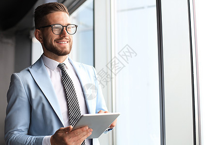 现代正装商务人士在办公室靠近窗口时使用数字平板电脑 同时站在办公室窗户附近阶层地点衬衫营销领带工作男性白领经理男人图片