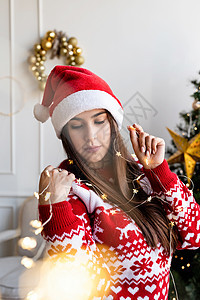 穿着圣塔帽和毛衣的年轻女子 带着美丽的圣诞光假期房子女性闲暇季节新年庆典成人礼物房间图片