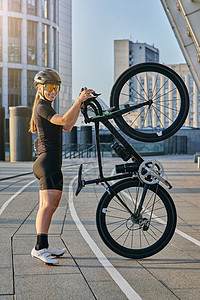 在阳光明媚的日子里 身着黑色自行车服和防护装备的职业女自行车手对着镜头微笑 她的自行车直立在户外摆姿势图片