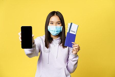 面戴面具 展示智能手机屏幕 应用程序接口和护照及假期票 预订旅行 黄色背景的韩国快乐女孩 并配有度假机票图片