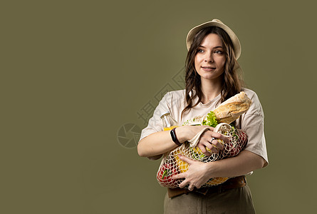 手插口袋的女孩穿着浅色夏装 带着装满蔬菜的网眼生态袋 在绿色工作室背景下的相机里看着绿色植物的年轻女子微笑着 可持续的生活方式 生态友好的概念背景